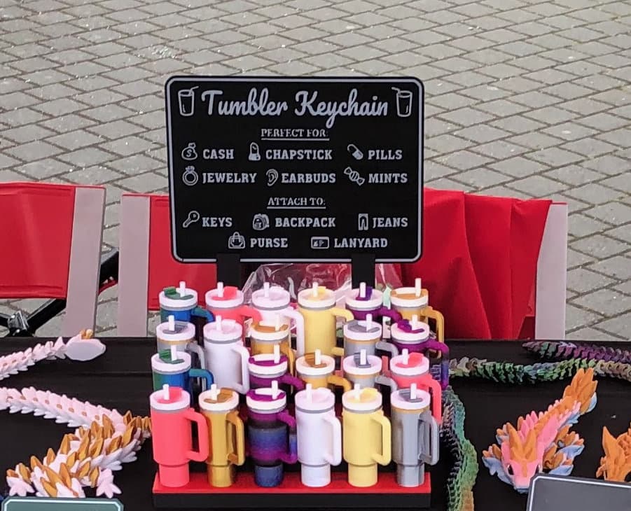 A tiered craft fair display of colorful 3D-printed mini tumbler keychain chapstick holders beneath a sign reading Tumbler Keychain.
