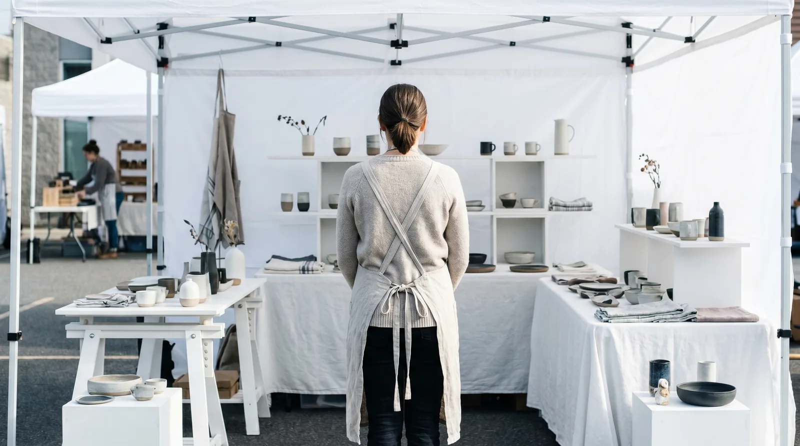 A vendor in a linen apron stands in front of her white-tented craft fair booth from the aisle perspective, taking a long look back at her ceramics display before the market opens