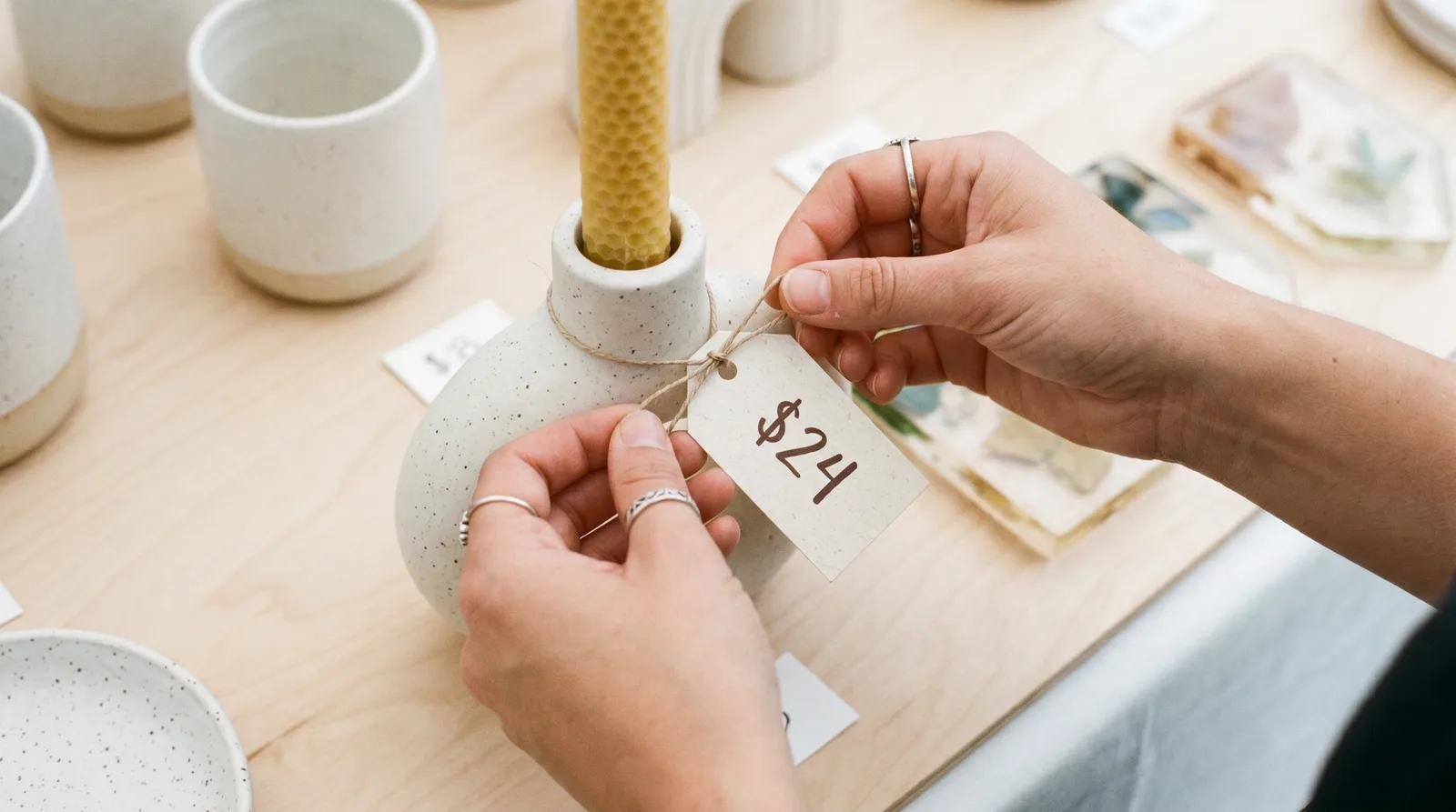 Close-up of a maker's hands tying a small kraft price tag reading "$24" onto a beeswax candle in a speckled ceramic holder, with other tagged items in soft focus across the table