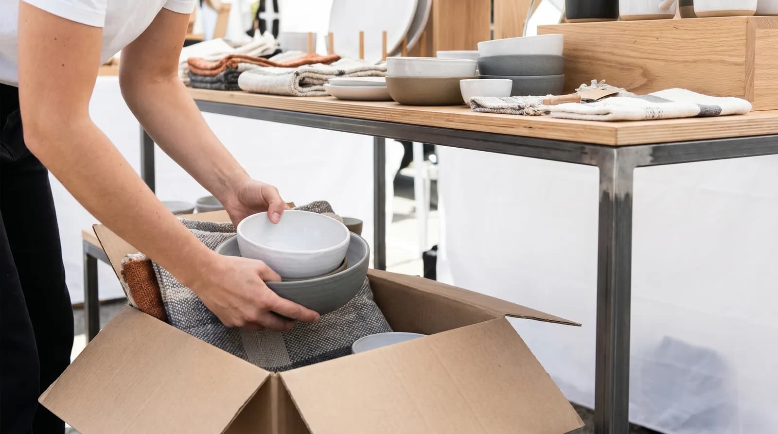 A vendor lifts stacked ceramic bowls from a labeled cardboard bin tucked beside her booth table, ready to restock the display without disrupting the front-of-table arrangement