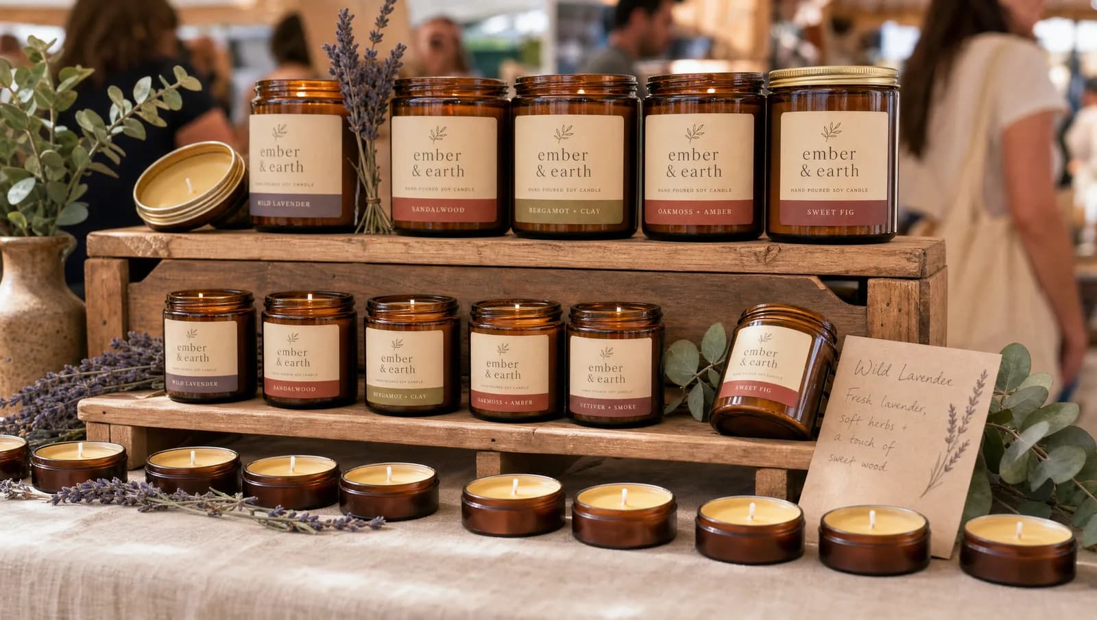 A candle vendor's booth with two tiers of amber jar candles on a wooden crate riser, smaller tealights lined up at the front edge of a linen-draped table, and lavender stems for visual softness