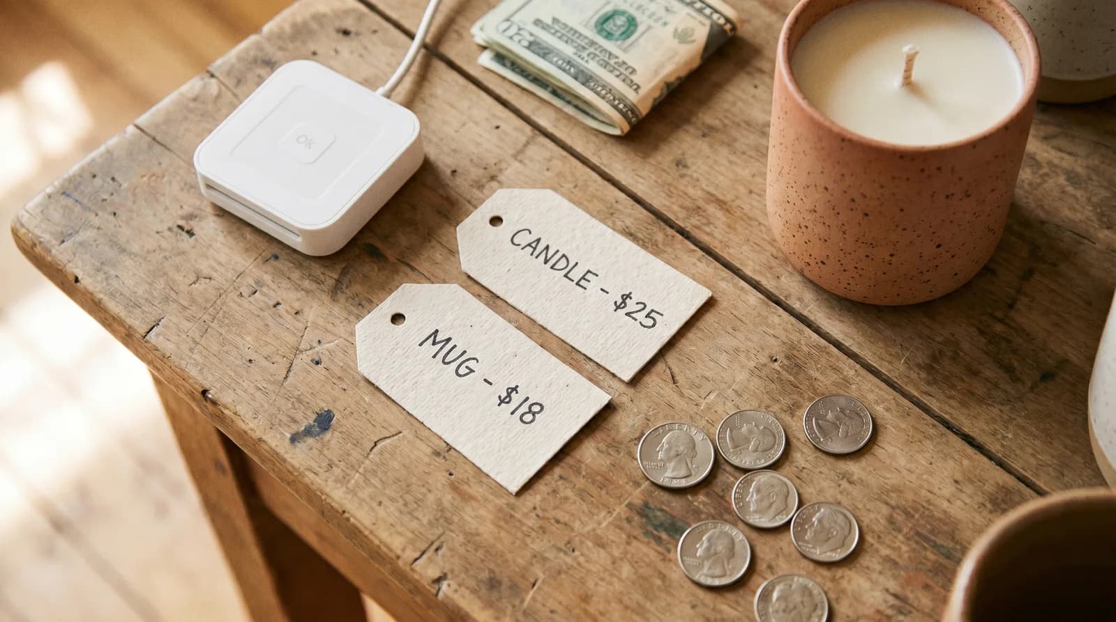 A Square card reader, a small stack of cash, and two handwritten price tags reading "Candle - $25" and "Mug - $18" on a wooden table, with a candle and coins nearby